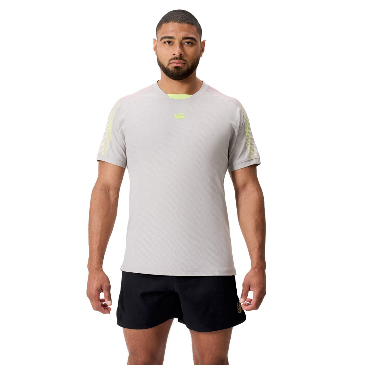 A man faces forward in a Canterbury Tempo+ Drill Top, light gray and made from moisture-wicking fabric, paired with black shorts against a plain white background.