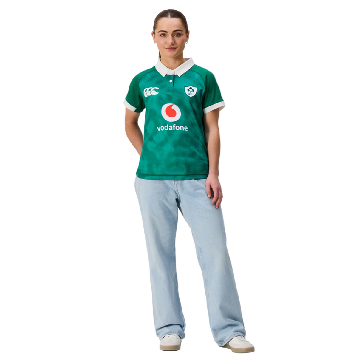 A person stands facing forward, wearing the Canterbury Women's Ireland 25/26 Home Pro Jersey with light blue jeans and white sneakers against a plain white background.