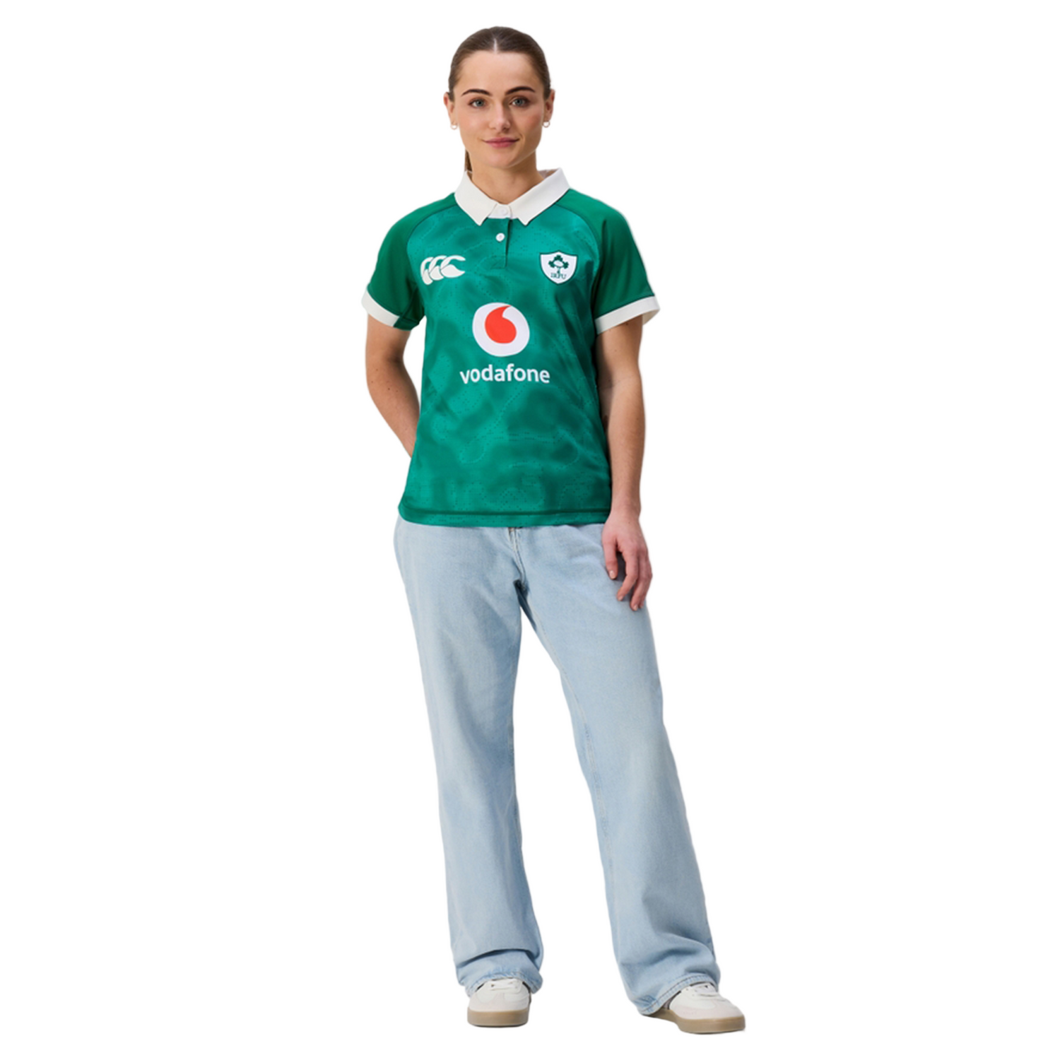 A person stands facing forward, wearing the Canterbury Women's Ireland 25/26 Home Pro Jersey with light blue jeans and white sneakers against a plain white background.