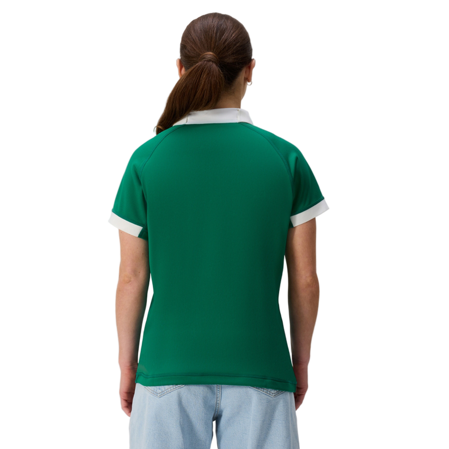 A woman with long hair in a ponytail is seen from the back wearing the Canterbury Women's Ireland 25/26 Home Pro Jersey with white trim and light blue jeans, standing against a plain background.