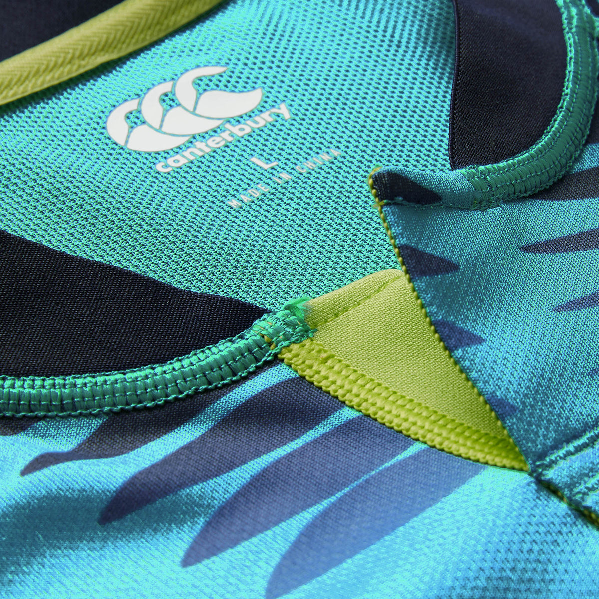 Close-up of the Ireland 26 Pre Game Pro Jersey by Canterbury in teal and black, highlighting the signature logo, size label, embroidered seams, and a section of striking printed pattern. This moisture-wicking Canterbury sports jersey combines detailed construction with high-performance style.
