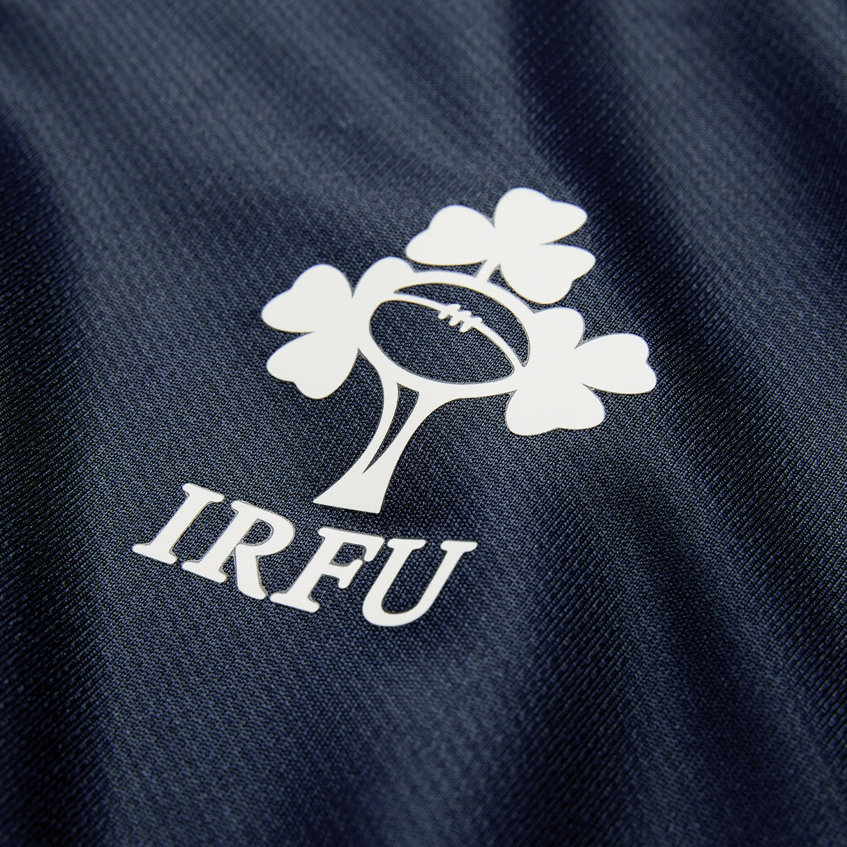 The Ireland 26 Training Pro Jersey by Canterbury features the IRFU logo—a shamrock and rugby ball design—printed in white on dark blue textured fabric, and incorporates Canterbury’s signature VapoDri technology for optimal comfort.