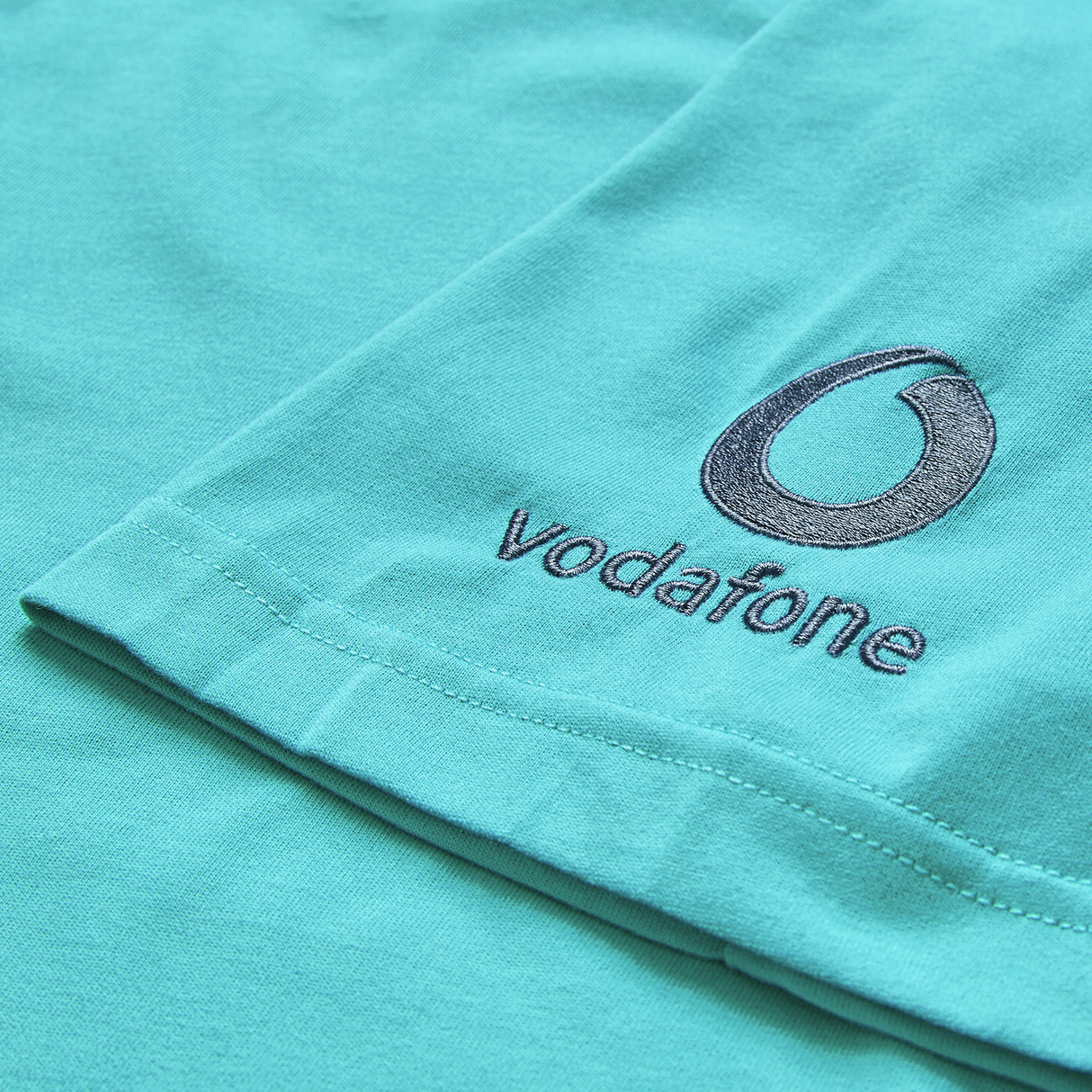Close-up of the Ireland 26/27 Summer Tour Team Cotton T-Shirt by Canterbury, featuring embroidered Vodafone logo and text on turquoise fabric.