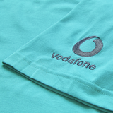 Close-up of the Ireland 26/27 Summer Tour Team Cotton T-Shirt by Canterbury, featuring embroidered Vodafone logo and text on turquoise fabric.