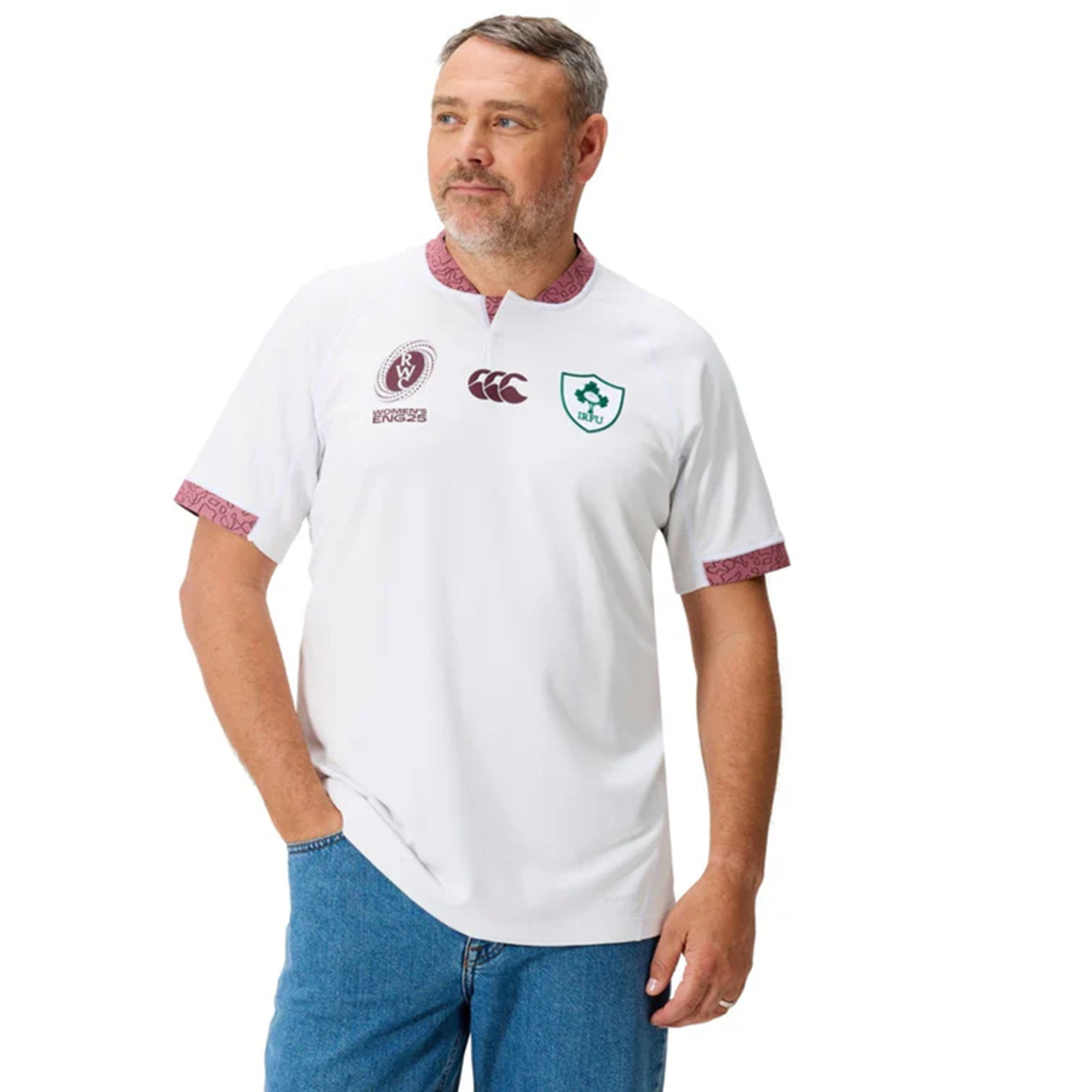 A man wearing the Ireland WRWC25 Replica Alternate Jersey by Canterbury, a white short-sleeve rugby shirt featuring maroon and green logos with patterned trim on the collar and sleeves, paired with blue jeans.
