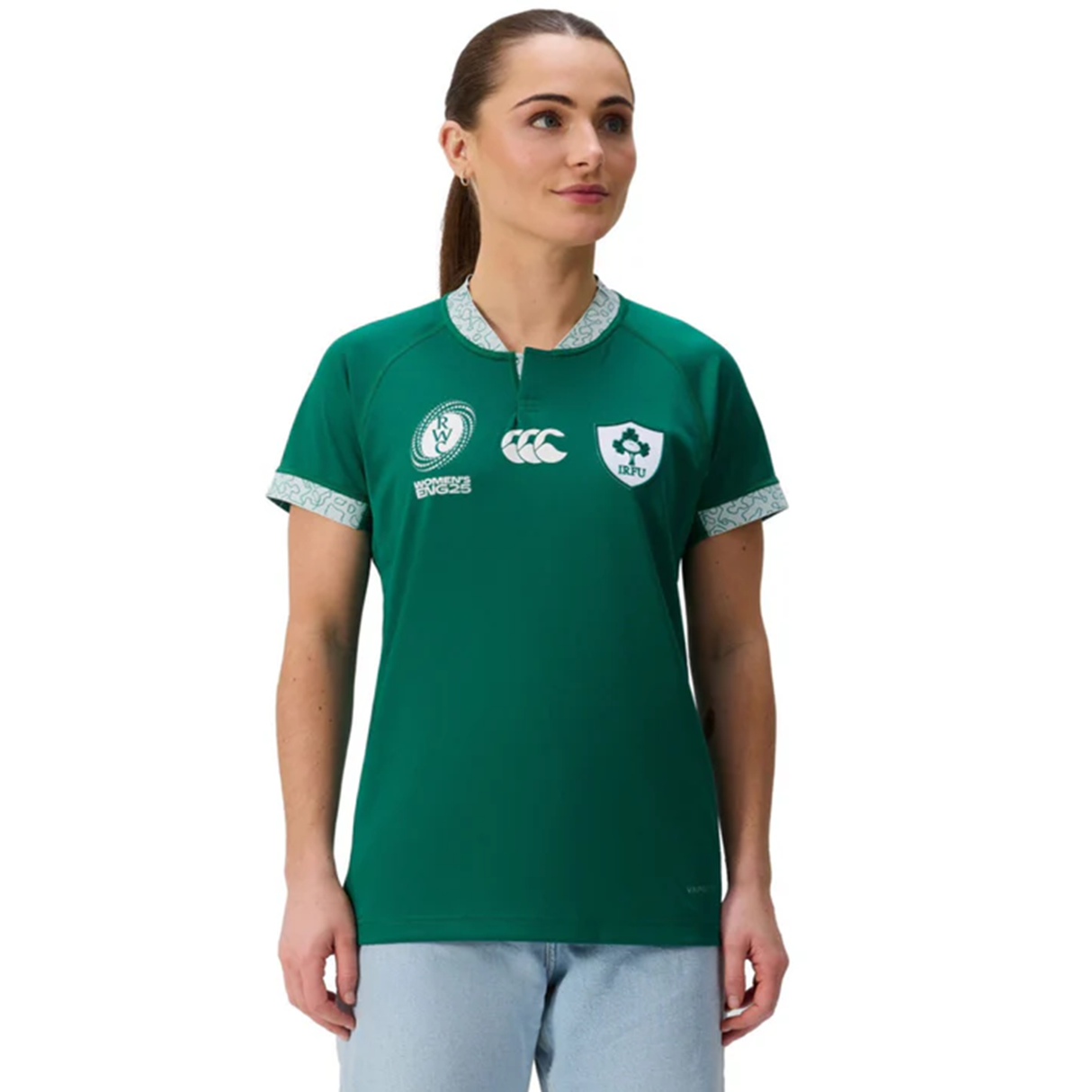 A woman wears the Canterbury Women's Ireland WRWC25 Replica Home Jersey with sponsor logos and light blue jeans, facing slightly left against a plain white background.