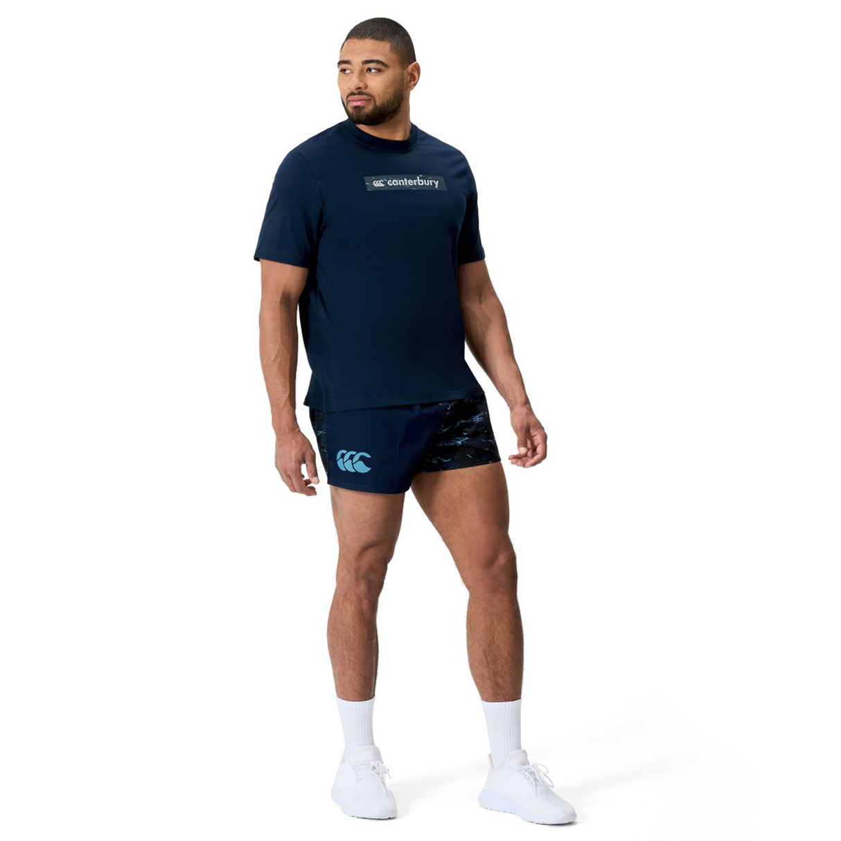 A man faces left against a white background, dressed in a navy Canterbury t-shirt, Force Harlequin Drill Shorts by Canterbury, white socks, and white sneakers.