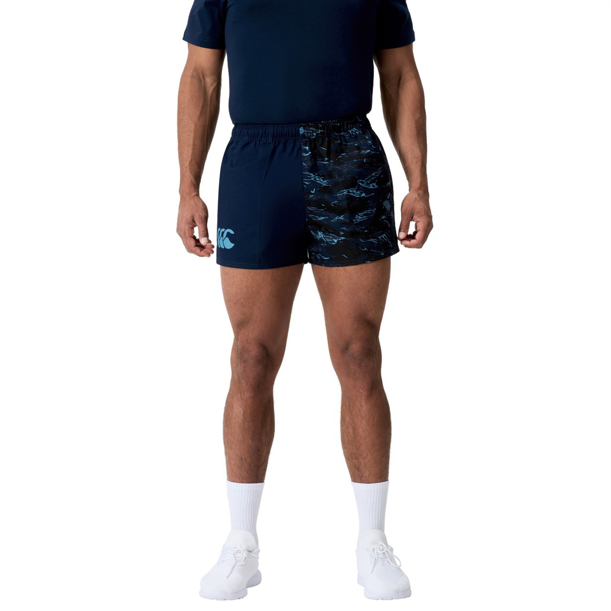 A person wearing a navy Canterbury athletic outfit with Force Harlequin Drill Shorts by Canterbury, white socks, and white sneakers stands against a plain background.
