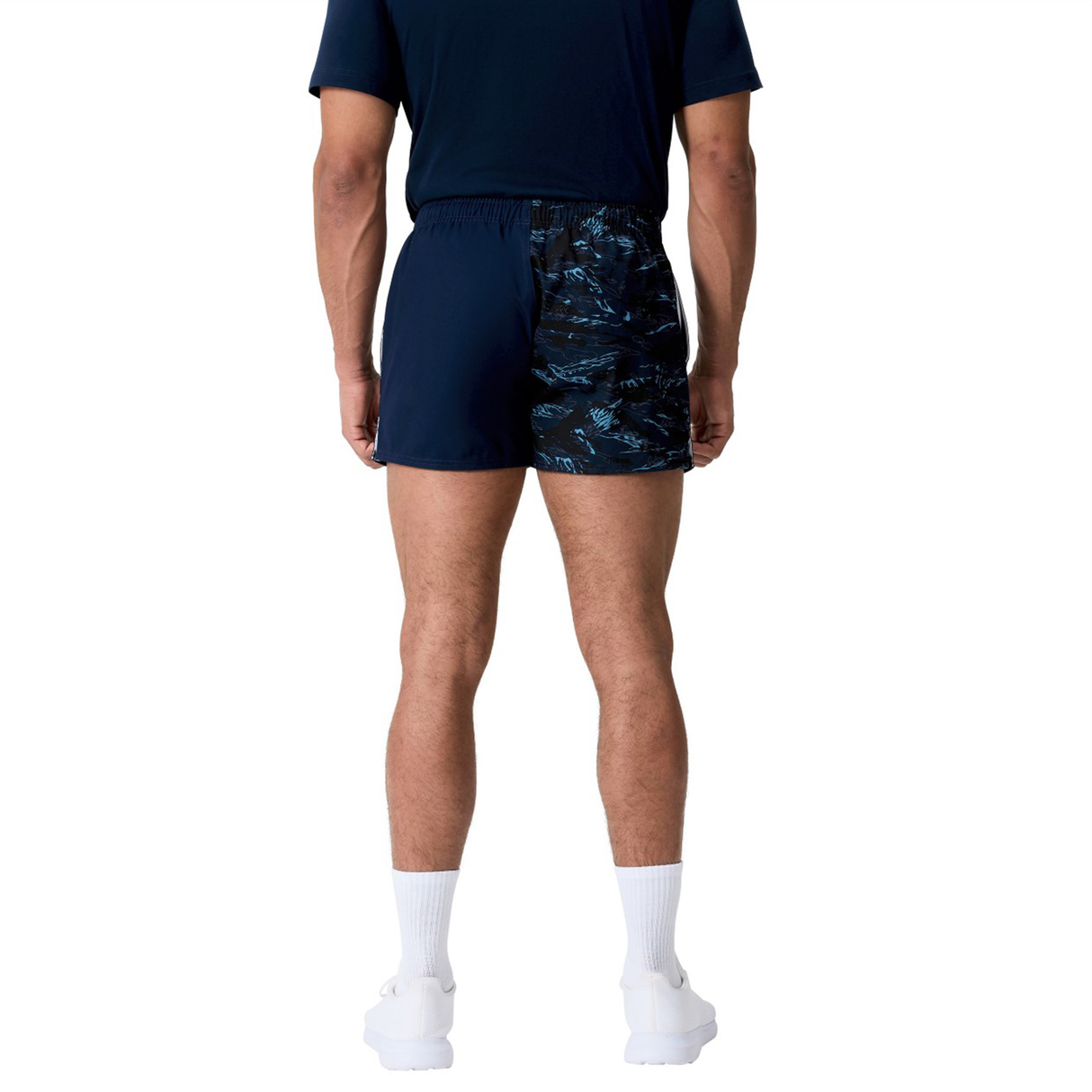 A man stands facing away against a plain white background, wearing a navy t-shirt, Canterbury Force Harlequin Drill Shorts, white socks, and white athletic shoes.