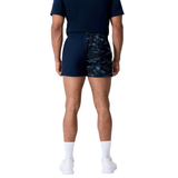A man stands facing away against a plain white background, wearing a navy t-shirt, Canterbury Force Harlequin Drill Shorts, white socks, and white athletic shoes.