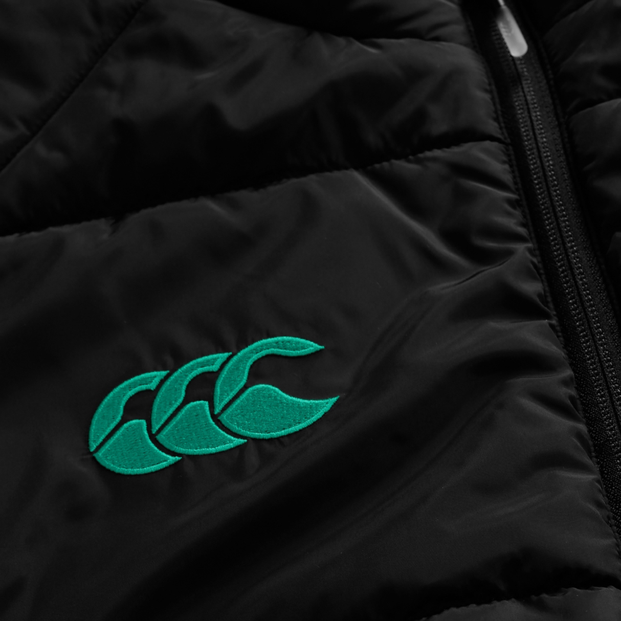 Close-up of the Ireland 25/26 Elite Gilet by Canterbury: a black gilet with a green embroidered logo of three overlapping shapes and a visible black zipper—ideal for Ireland rugby fans.
