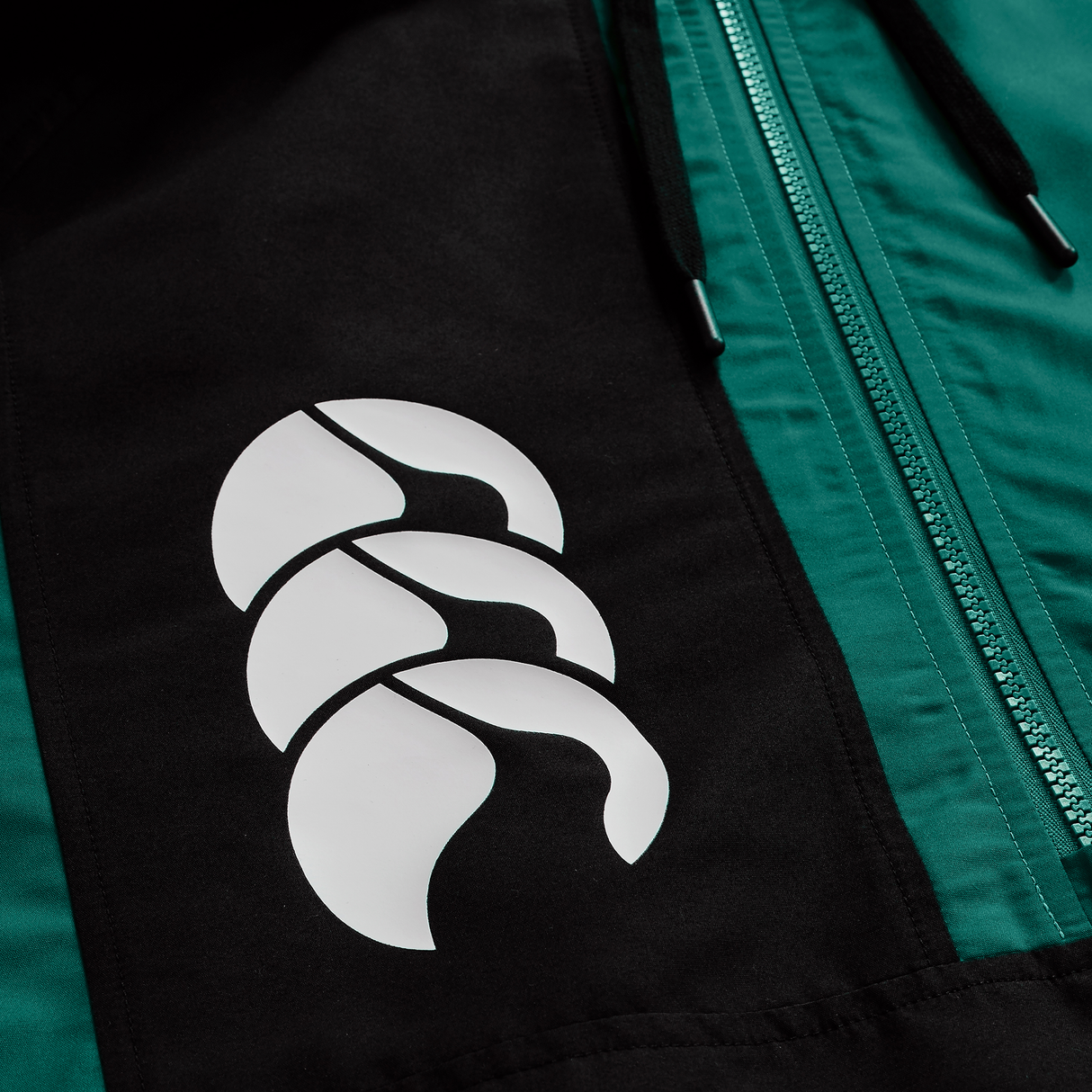 A close-up of the Ireland 25/26 Legends 1/4 Zip Jacket by Canterbury features a white abstract logo and visible green zipper, emphasizing the brand's stylish quarter-zip design.