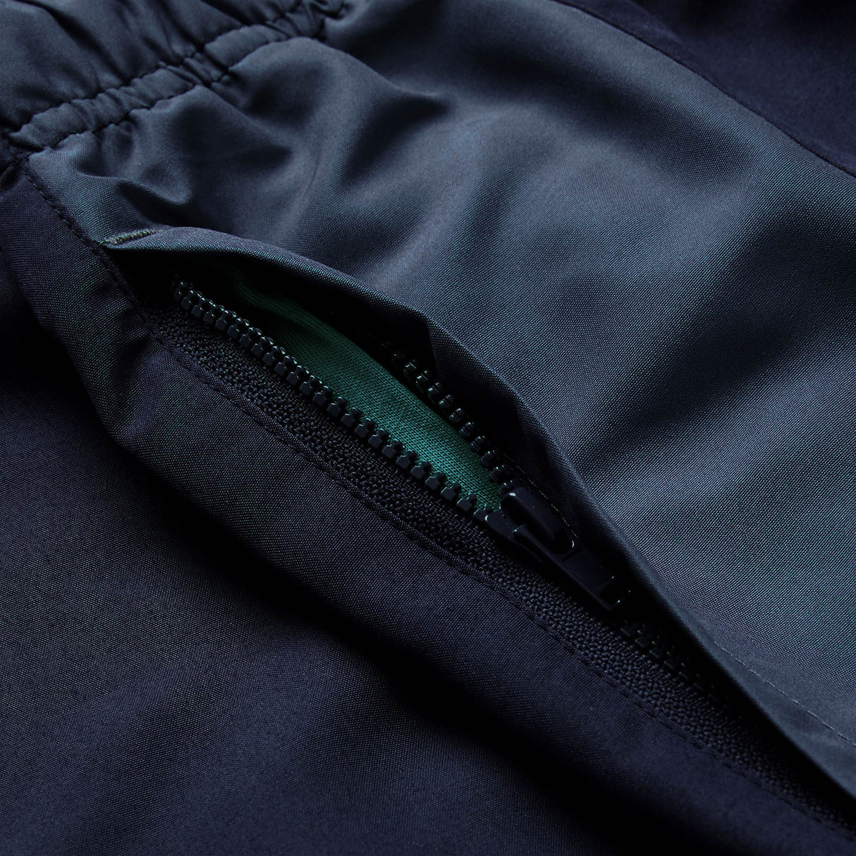 Close-up of a dark-colored garment with an open zipper pocket, revealing green fabric inside and an elastic waistband at the top—features that showcase the quality construction typical of Canterbury’s Ireland 26 Legends 32″ Trackpant.