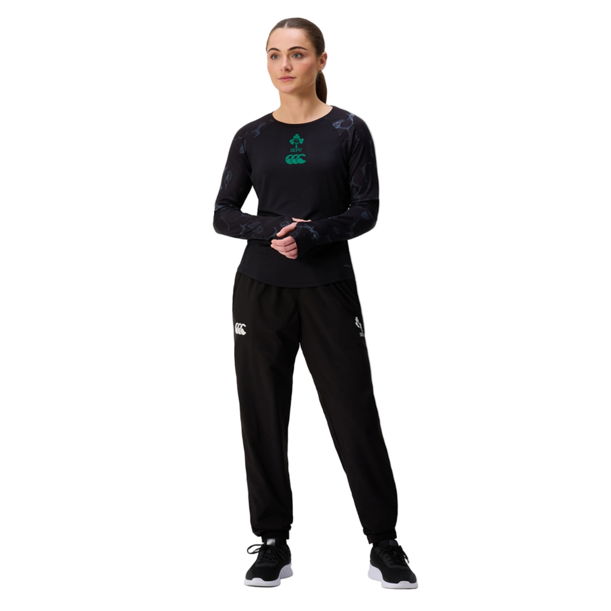 A woman stands against a white background wearing a black long-sleeve shirt, black sneakers, and Canterbury Women's Ireland 25/26 Training Pants featuring branded logos.