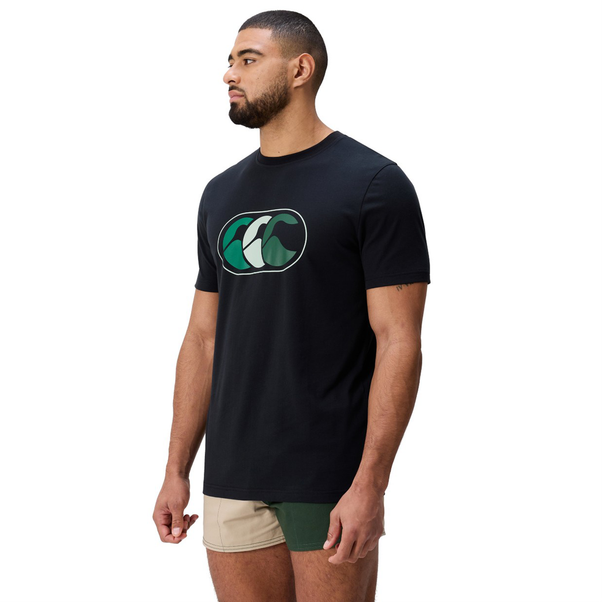 A bearded man stands looking left, wearing the FUN. T-SHIRT AM and Canterbury's two-tone green and beige shorts with a green wave logo.