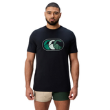 A man stands facing forward, wearing the Canterbury FUN. T-SHIRT AM with a stylized green wave logo and two-tone shorts—one side beige, the other dark green.
