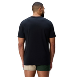 A person stands facing away from the camera, wearing a black Canterbury FUN. T-SHIRT AM and color-blocked shorts with tan and green panels.