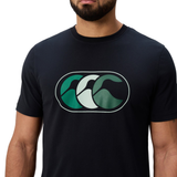 A man wears the Canterbury FUN. T-SHIRT AM, featuring a stylized green and blue wave logo on the chest, photographed against a white background.