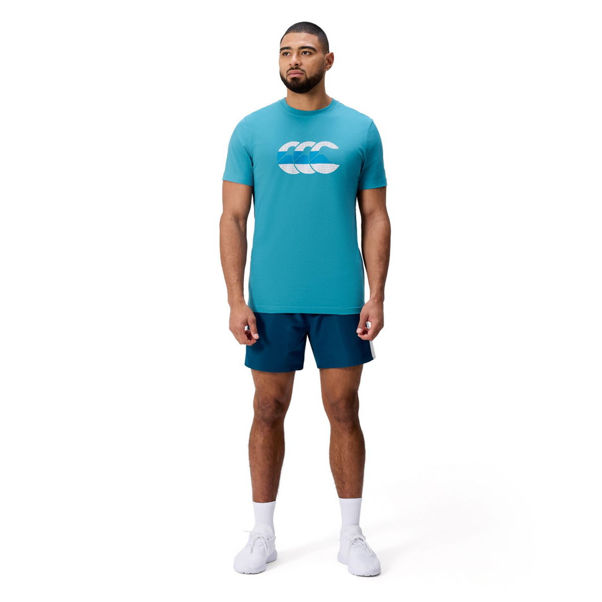 A man stands facing forward wearing a light blue Canterbury FUN. T-SHIRT AM with a white logo, dark blue shorts, white socks, and white athletic shoes against a plain white background.