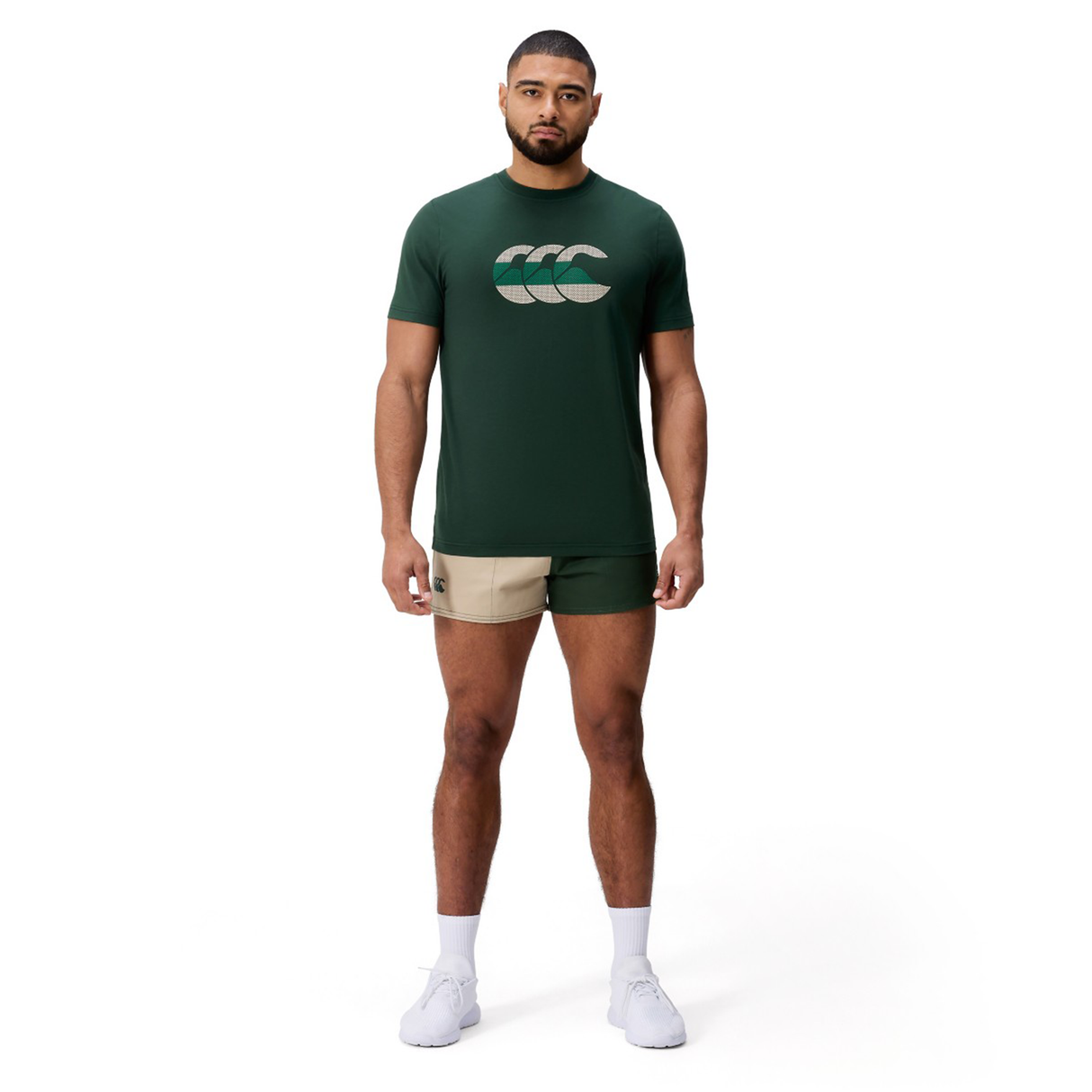 A man faces forward against a white background, wearing a dark green Canterbury FUN. T-SHIRT AM with logo, beige athletic shorts, white socks, and white shoes.