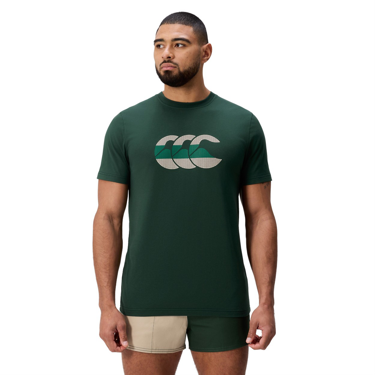 A man faces forward, dressed in a dark green Canterbury FUN. T-SHIRT AM featuring a geometric logo and beige-green shorts, standing against a plain white background.