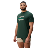 A man stands at an angle against a white background, wearing a Canterbury FUN. T-SHIRT AM in dark green with a bold logo and beige-green shorts.