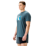 A man wearing the Canterbury FUN. T-SHIRT AM, a teal graphic t-shirt with a wave logo, and black athletic shorts stands facing left against a plain white background.