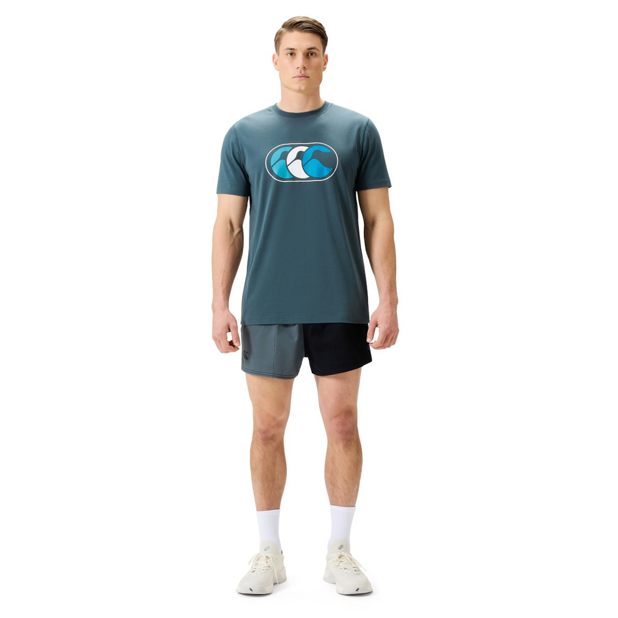 A man faces forward against a plain white background, wearing a Canterbury FUN. T-SHIRT AM in blue with black and gray shorts, white socks, and white sneakers.