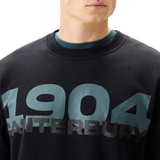 A person wearing Canterbury’s EVERYDAY GFX CREW SWEATER in black, featuring bold blue and gray "1904 CANTERBURY" lettering across the chest—a stylish choice for daily wear.