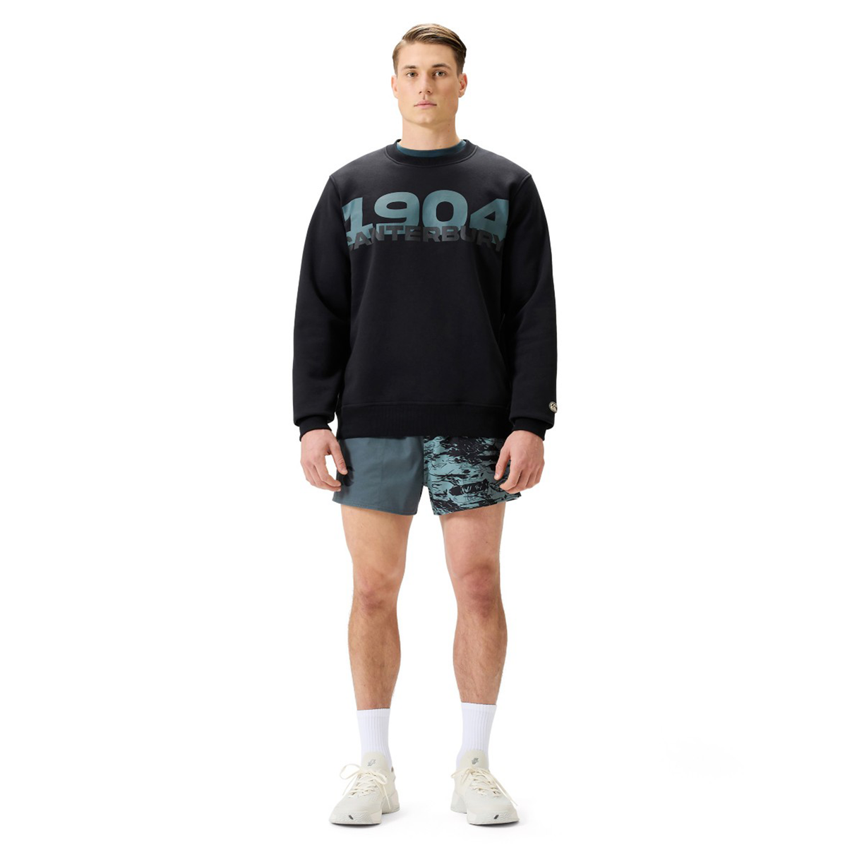 A man poses against a white background wearing the Canterbury EVERYDAY GFX CREW SWEATER with "1904" on the front, paired with blue patterned shorts, white socks, and sneakers—a comfortable look for daily wear.