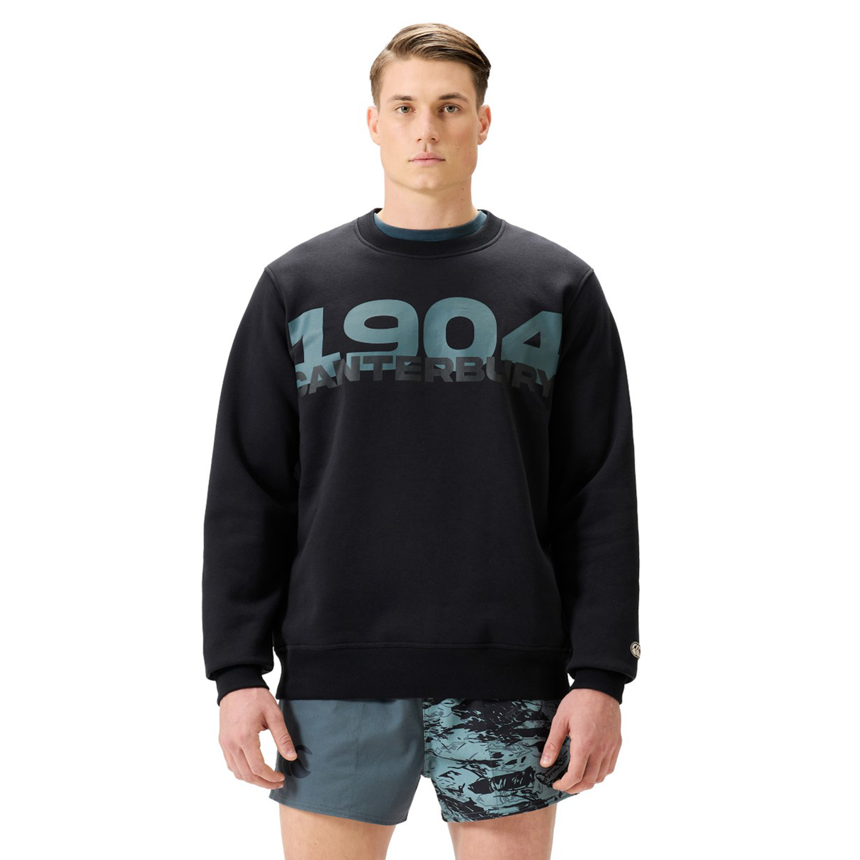 A man wears a black Canterbury EVERYDAY GFX CREW SWEATER with patterned blue shorts, standing against a plain white background—ideal for daily wear.