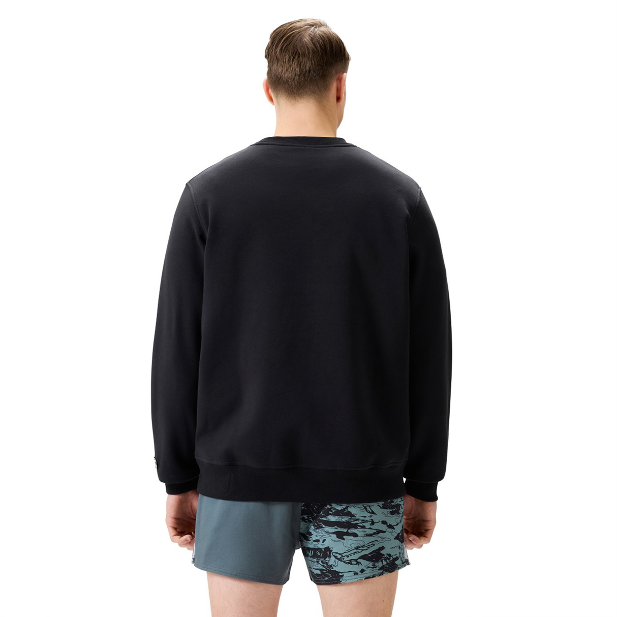 A man, facing away from the camera, wears a Canterbury EVERYDAY GFX CREW SWEATER in black with blue patterned shorts—ideal everyday wear against a plain white background.