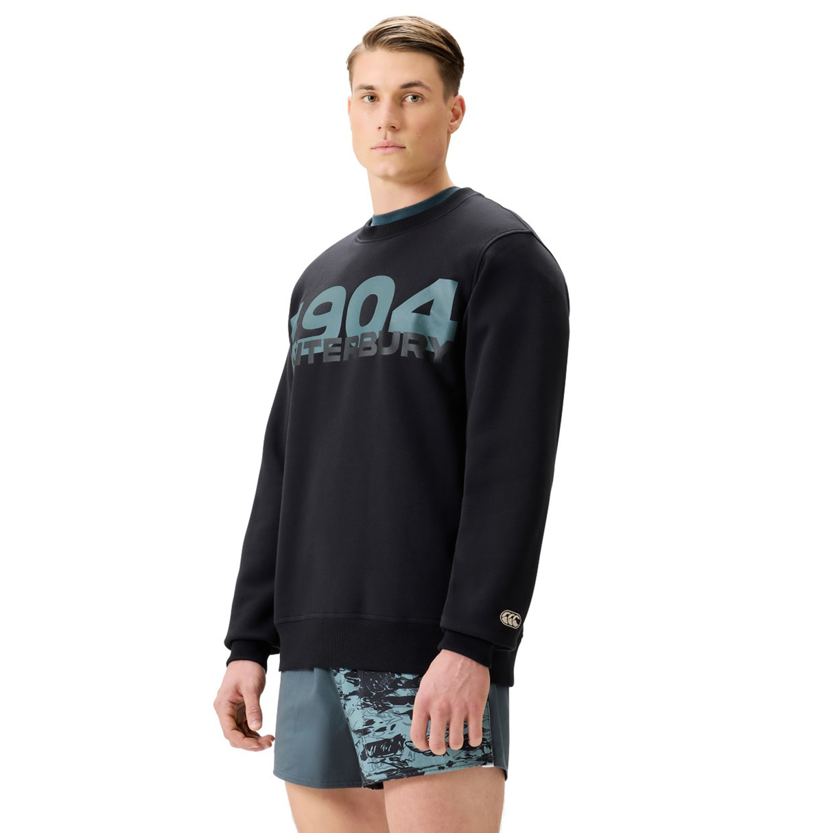 A man stands facing forward in a black Canterbury EVERYDAY GFX CREW SWEATER with "1904 Canterbury" on the front and patterned athletic shorts, showcasing a classic everyday look from Canterbury.