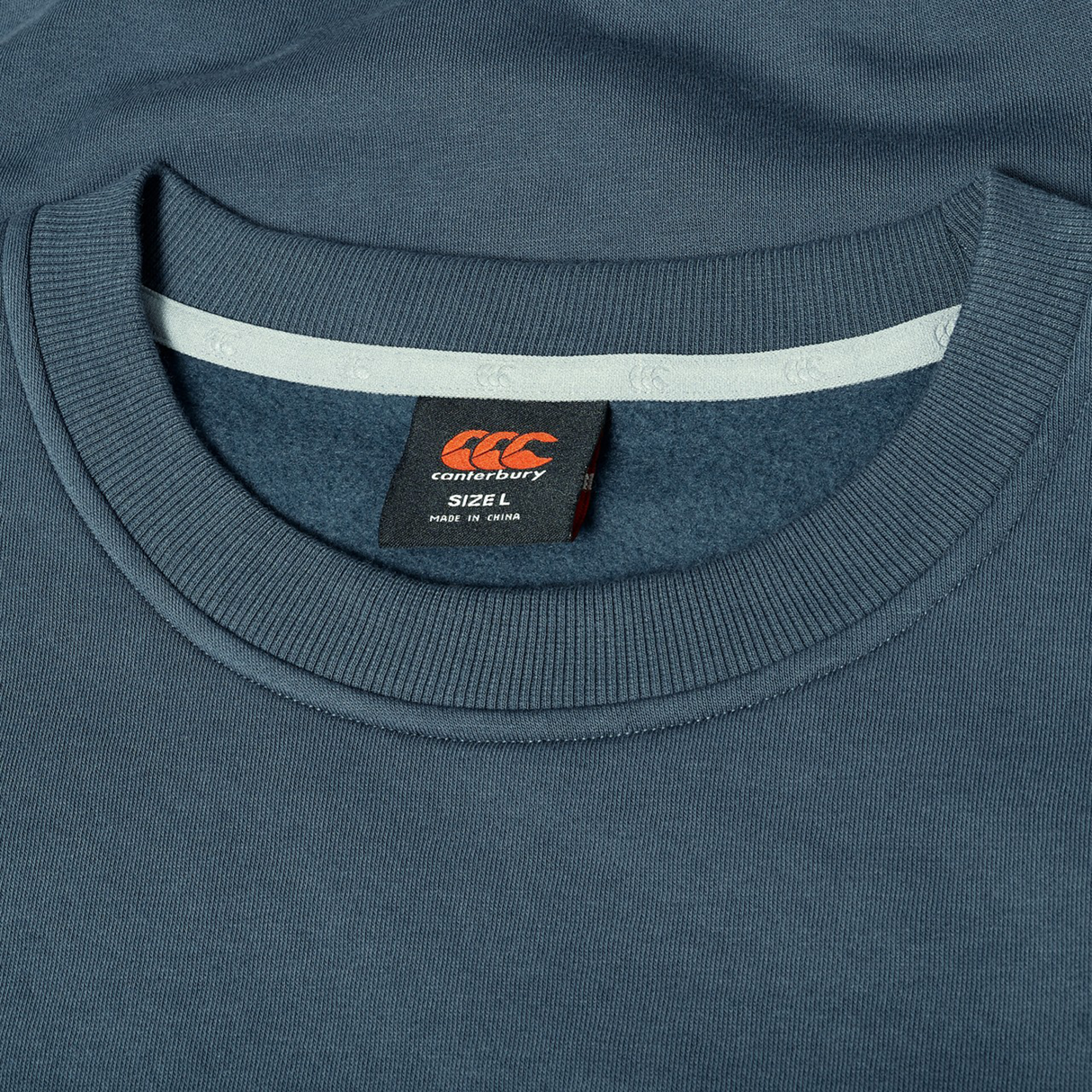 Close-up of a blue Canterbury EVERYDAY GFX CREW SWEATER laid flat, highlighting the collar and black size L label with orange logo and text—ideal for everyday wear.