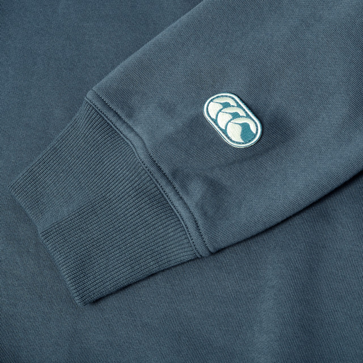 A close-up of the Canterbury EVERYDAY GFX CREW SWEATER’s blue cuff and sleeve, showing a small embroidered logo with two white shapes in an oval—ideal for stylish everyday wear.