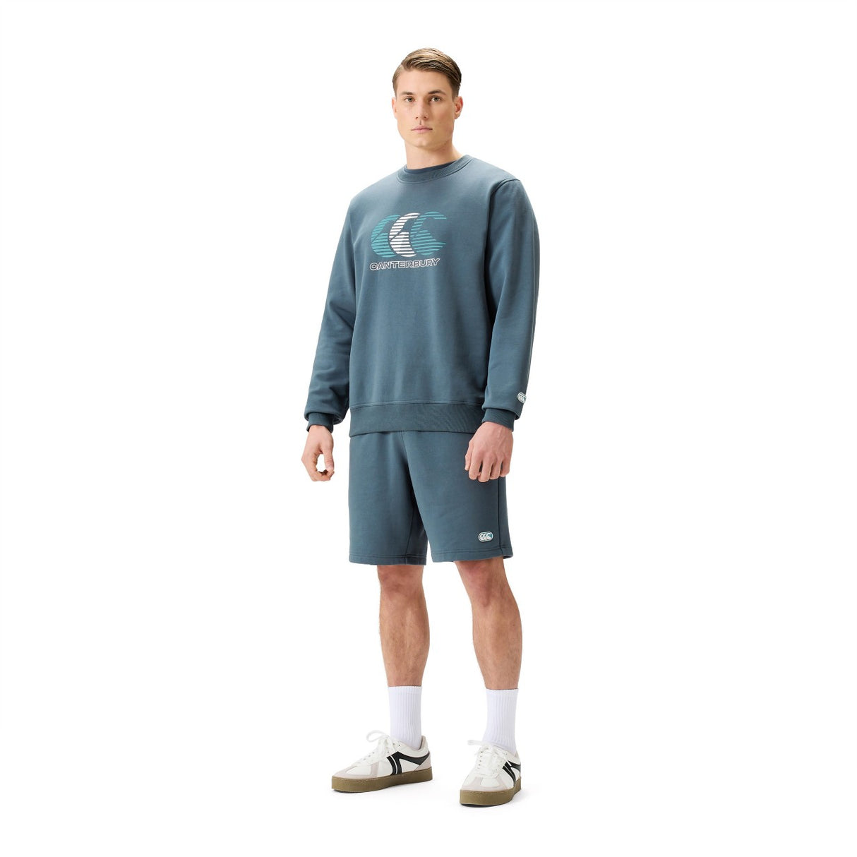 A man stands against a white background wearing Canterbury’s EVERYDAY GFX CREW SWEATER with a wave graphic, matching blue shorts, white socks, and beige sneakers—ideal for everyday style.