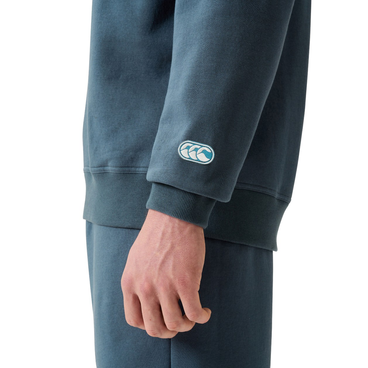 Someone wearing Canterbury’s EVERYDAY GFX CREW SWEATER in blue, featuring a white embroidered sleeve logo—ideal for daily wear and effortlessly matched with blue pants.