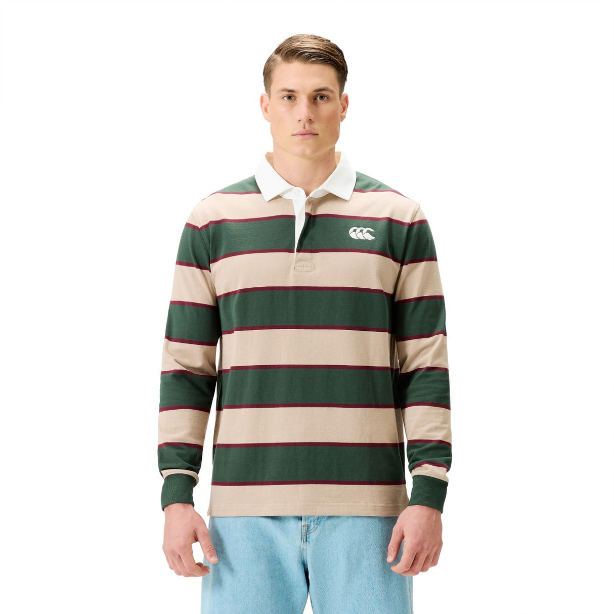 A man wears the Canterbury Stripe Rugby Jersey by Canterbury in green, beige, and red with a white collar, paired with blue jeans, standing against a plain white background.