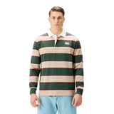 A man wears the Canterbury Stripe Rugby Jersey by Canterbury in green, beige, and red with a white collar, paired with blue jeans, standing against a plain white background.