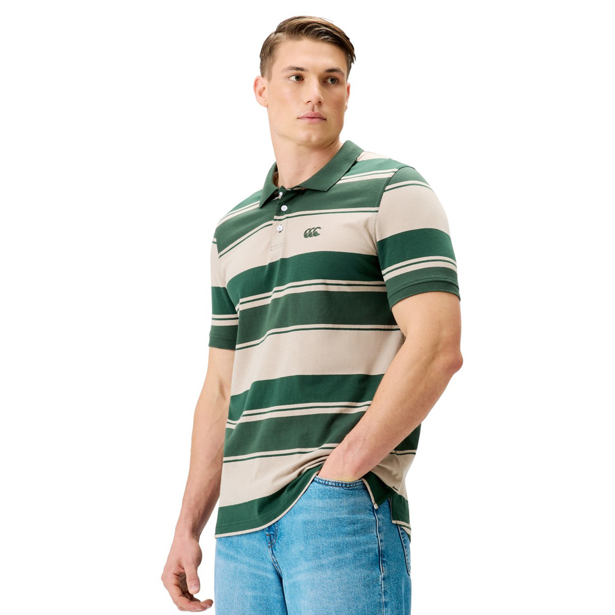A man in a Canterbury Yarn Stripe Polo by Canterbury, featuring green and beige stripes, and blue jeans stands against a white background, highlighting classic rugby style.