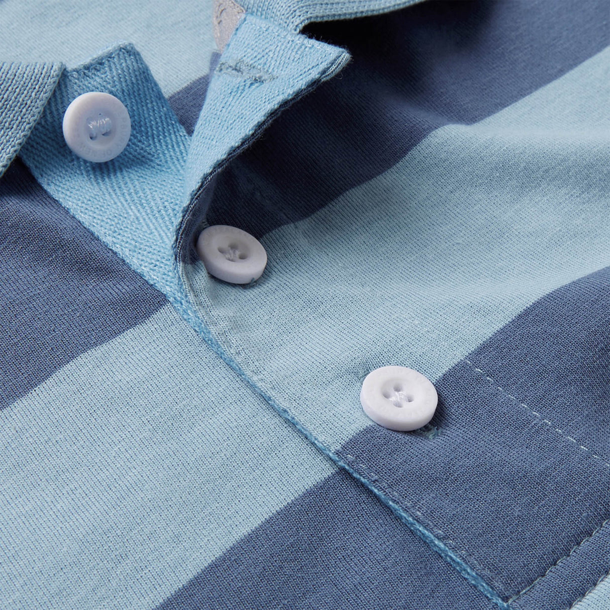 Close-up of the Canterbury Yarn Stripe Polo by Canterbury, featuring light blue and navy stripes and three white front placket buttons that highlight its classic rugby-inspired style.