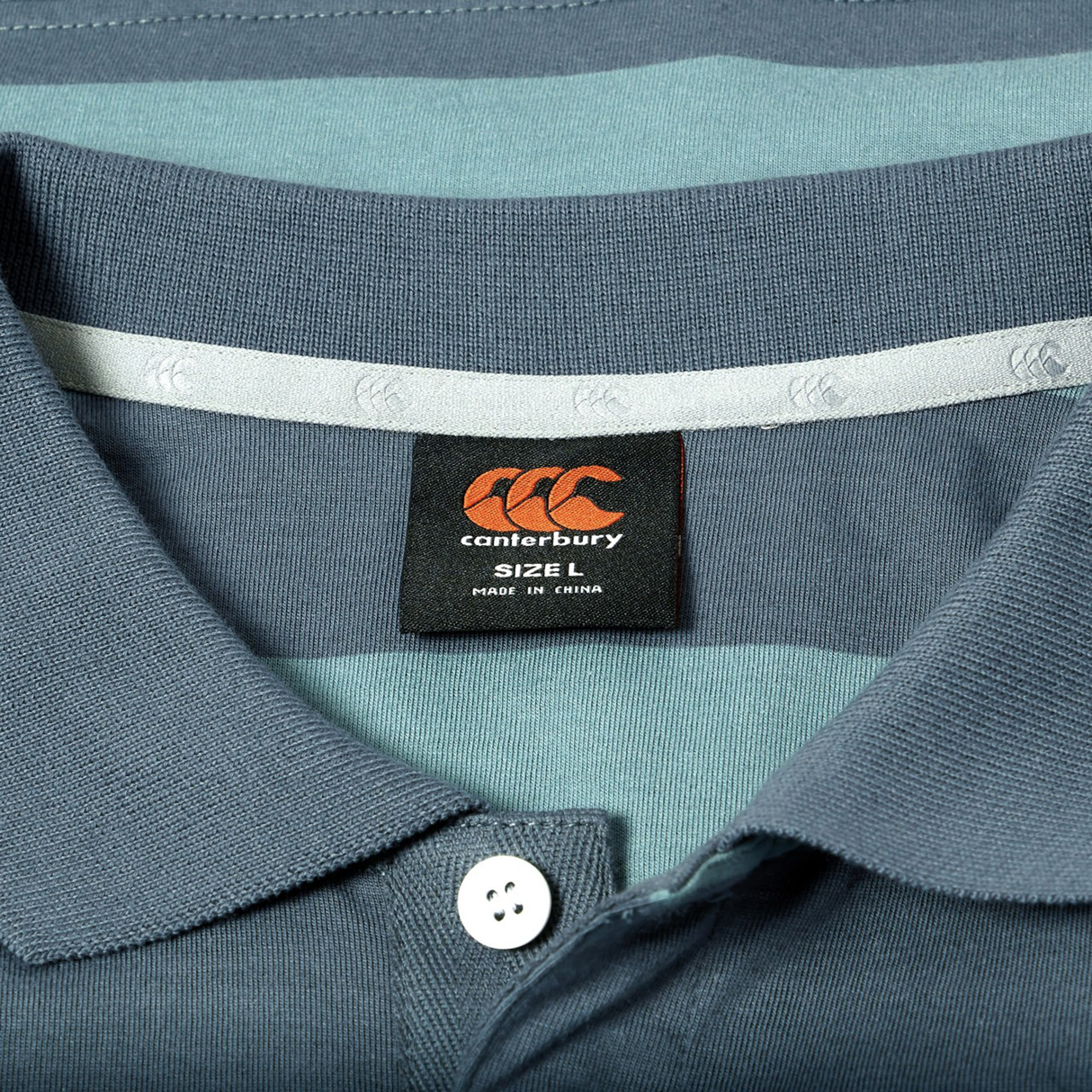 Close-up of the Canterbury Yarn Stripe Polo in blue and teal, showcasing classic rugby style details with a Canterbury brand label marked size L and Made in China under the collar.