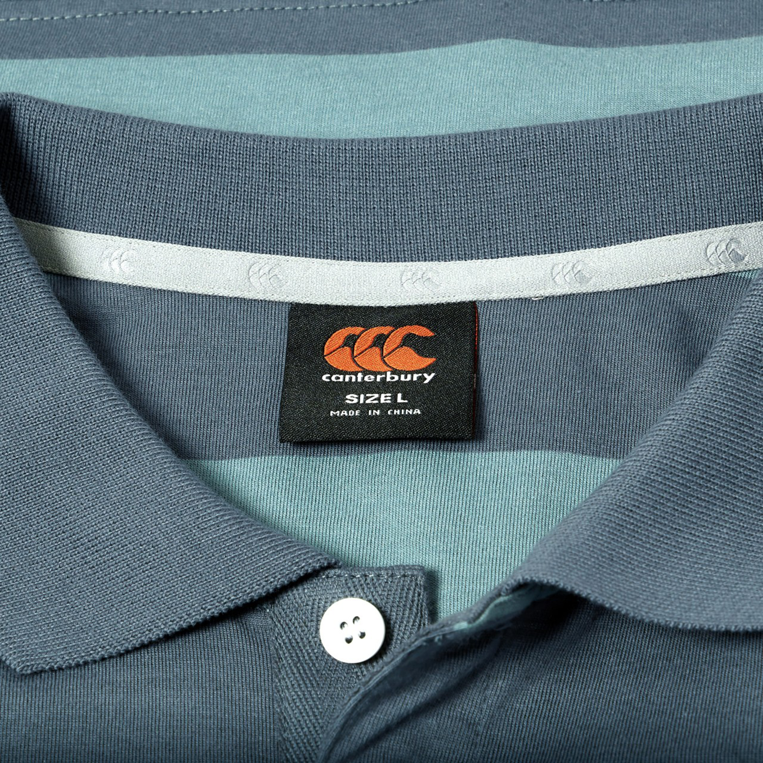 Close-up of the Canterbury Yarn Stripe Polo in blue and teal, showcasing classic rugby style details with a Canterbury brand label marked size L and Made in China under the collar.