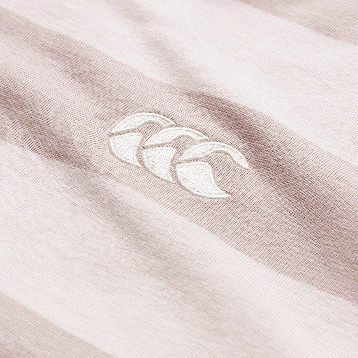 Here’s a rewritten version using the given product data: Close-up of a beige and white striped fabric from the Canterbury Yarn Stripe Polo by Canterbury, showcasing an embroidered logo with three overlapping curves in white thread, emphasizing its classic rugby-inspired style.