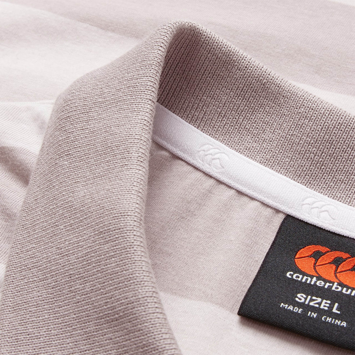 Close-up of the Canterbury Yarn Stripe Polo shirt collar in a light hue, featuring the Canterbury brand label, size L, and “Made in China” text—emphasizing its classic rugby-inspired style.