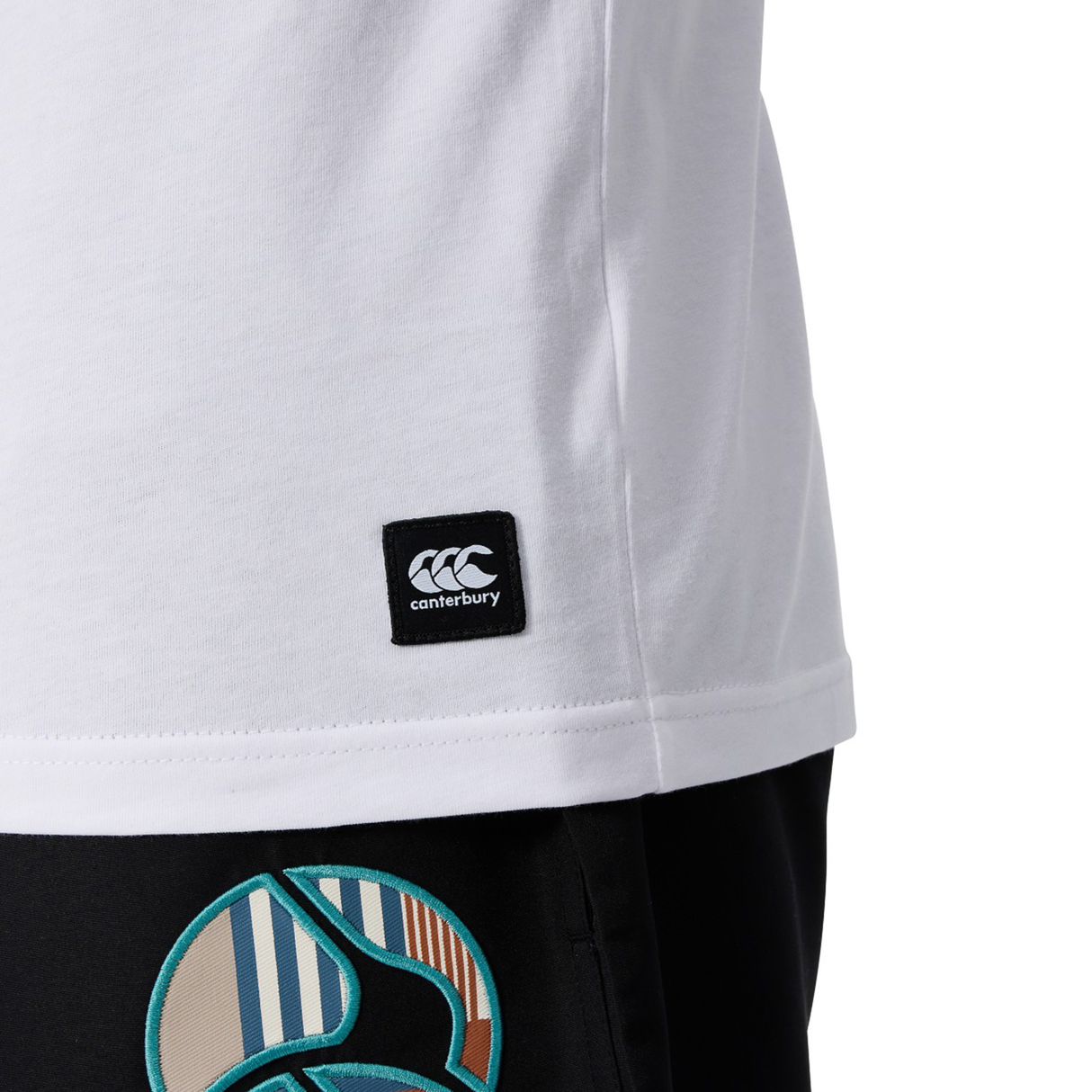 Close-up of a person in a white Canterbury Uglies Logo T-shirt by Canterbury, featuring a visible logo tag, paired with black pants with a colorful circular pattern.