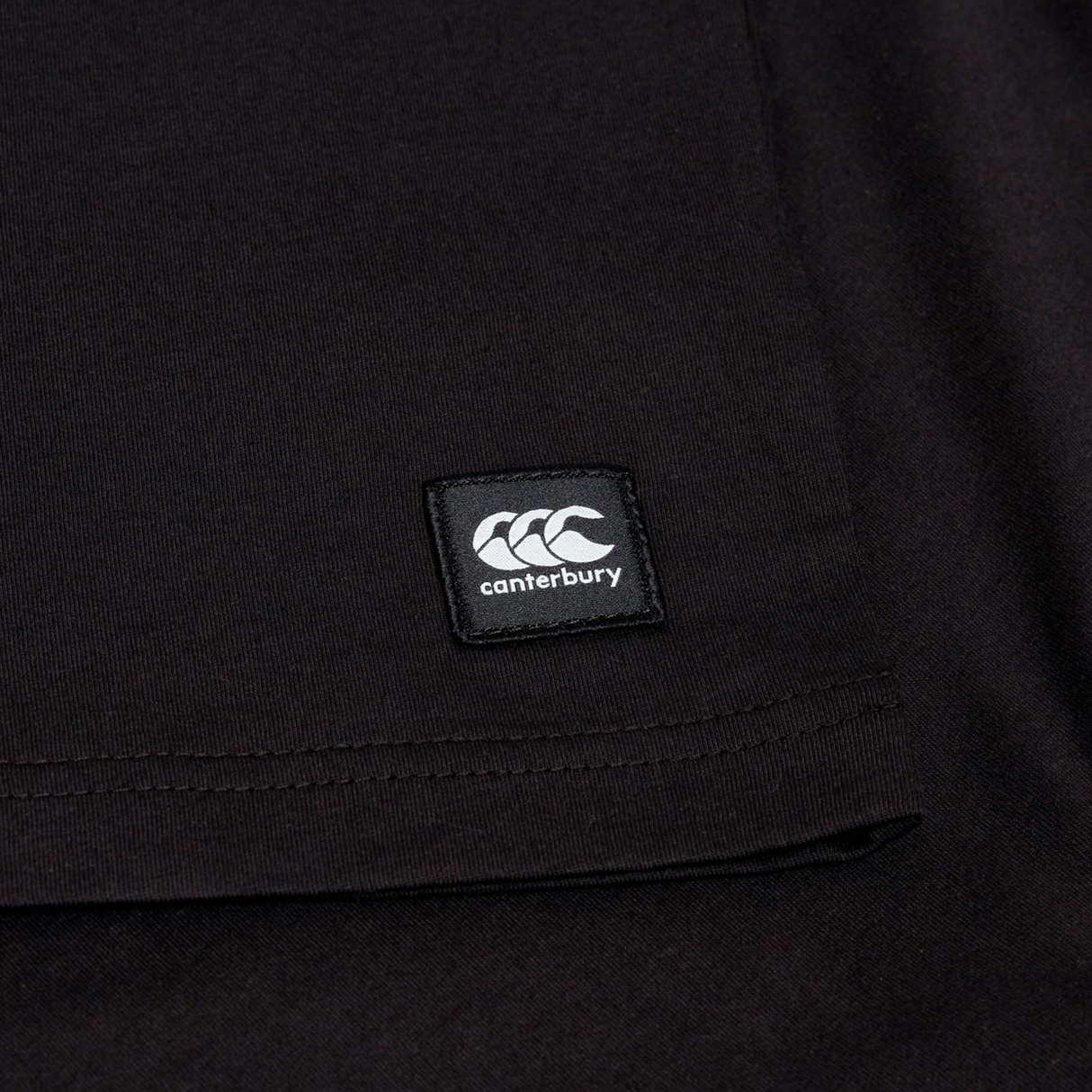 Close-up of the Canterbury Uglies Logo T-shirt in a black cotton blend, featuring a stitched rectangular Canterbury logo patch near the hem, inspired by classic Canterbury rugby shirts.