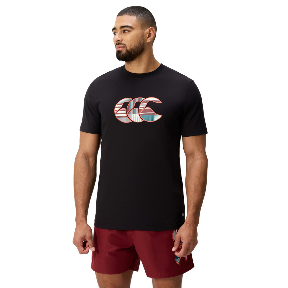 A bearded man wears a black Canterbury Uglies Logo T-shirt—a cotton blend tee with a patterned logo—and maroon athletic shorts, standing against a white background.