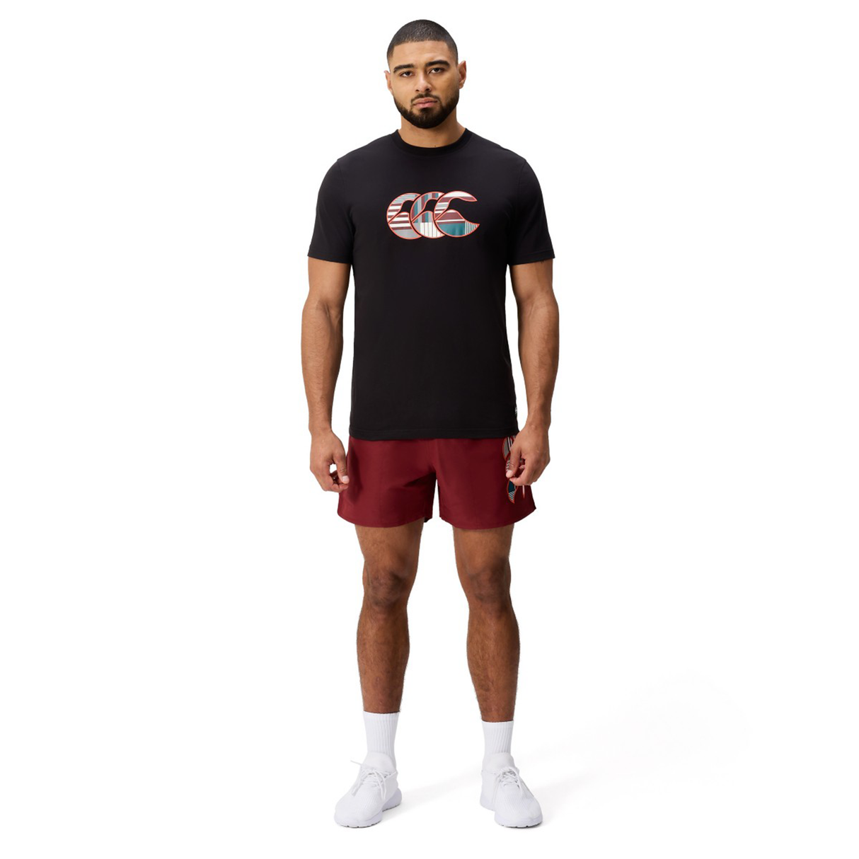 A man faces forward against a plain white background, wearing a black Canterbury Uglies Logo T-shirt with a patterned logo, maroon shorts, white socks, and white athletic shoes.