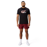 A man faces forward against a plain white background, wearing a black Canterbury Uglies Logo T-shirt with a patterned logo, maroon shorts, white socks, and white athletic shoes.